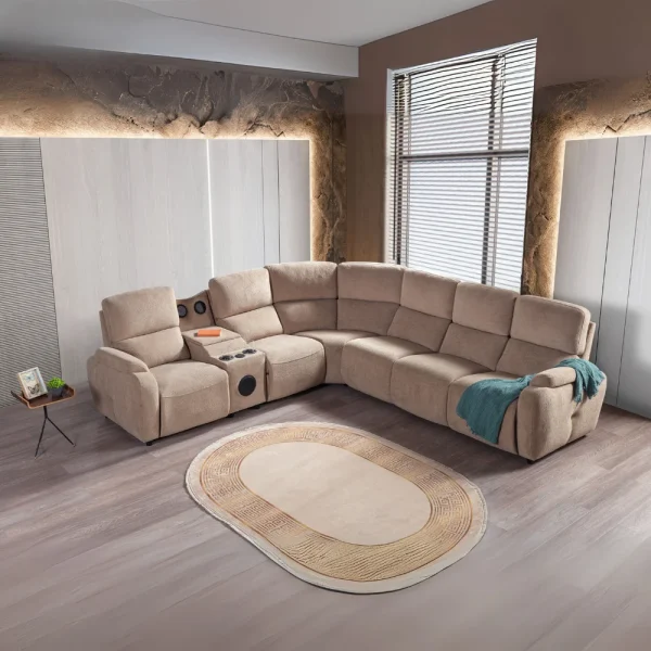 A detailed view of the Atlas Recliner Köşe Takımı, a contemporary L-shaped corner sectional sofa upholstered in textured beige fabric. The image shows its modern design with sleek lines and wide seats. The central console unit is featured, with an open lid, integrated cup holders, and a built-in Bluetooth speaker system visible on the side. The sofa is placed in a premium living room with light wood floors and textured wall paneling with LED backlighting.