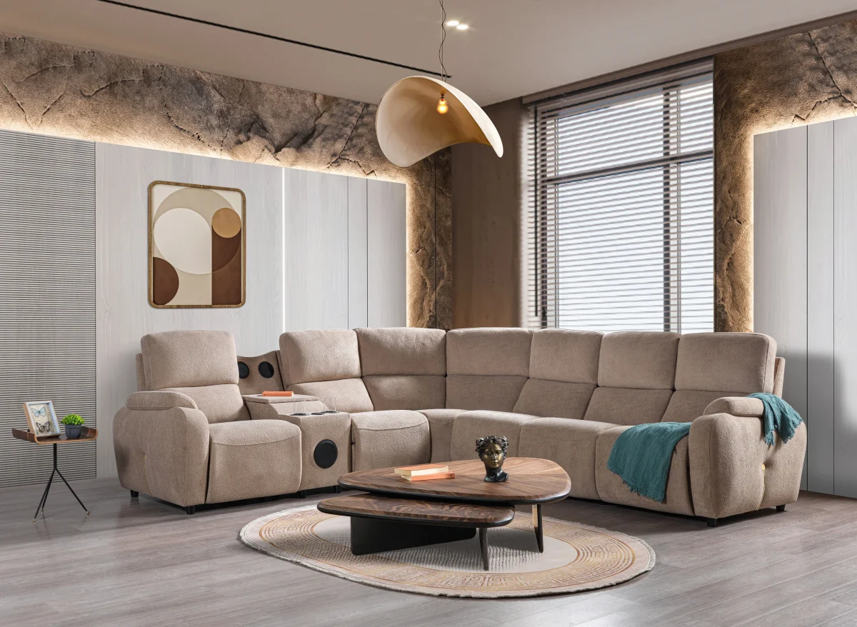 A detailed view of the Atlas Recliner Köşe Takımı, a contemporary L-shaped corner sectional sofa upholstered in textured beige fabric. The image shows its modern design with sleek lines and wide seats. The central console unit is featured, with an open lid, integrated cup holders, and a built-in Bluetooth speaker system visible on the side. The sofa is placed in a premium living room with light wood floors and textured wall paneling with LED backlighting.