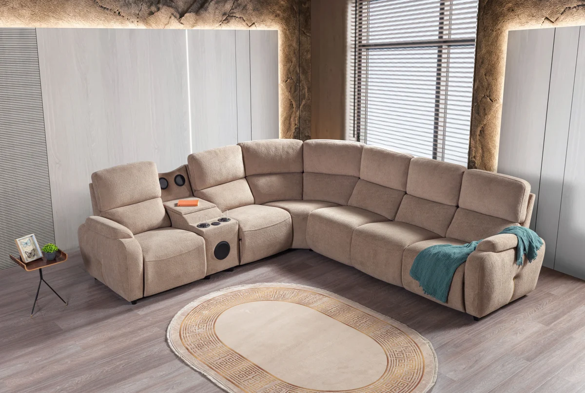 A detailed view of the Atlas Recliner Köşe Takımı, a contemporary L-shaped corner sectional sofa upholstered in textured beige fabric. The image shows its modern design with sleek lines and wide seats. The central console unit is featured, with an open lid, integrated cup holders, and a built-in Bluetooth speaker system visible on the side. The sofa is placed in a premium living room with light wood floors and textured wall paneling with LED backlighting.