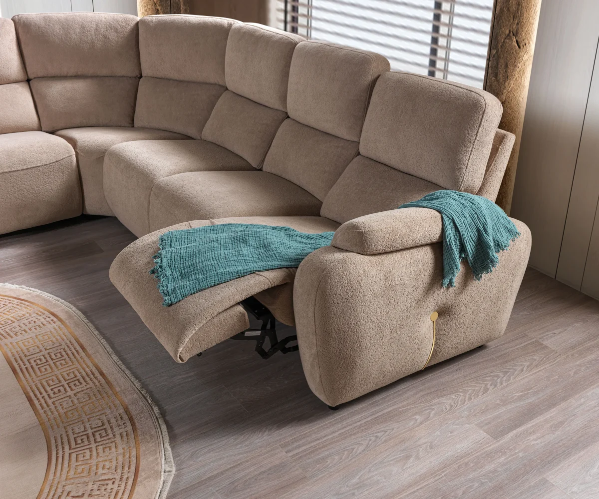 A detailed view of the Atlas Recliner Köşe Takımı, a contemporary L-shaped corner sectional sofa upholstered in textured beige fabric. The image shows its modern design with sleek lines and wide seats. The central console unit is featured, with an open lid, integrated cup holders, and a built-in Bluetooth speaker system visible on the side. The sofa is placed in a premium living room with light wood floors and textured wall paneling with LED backlighting.