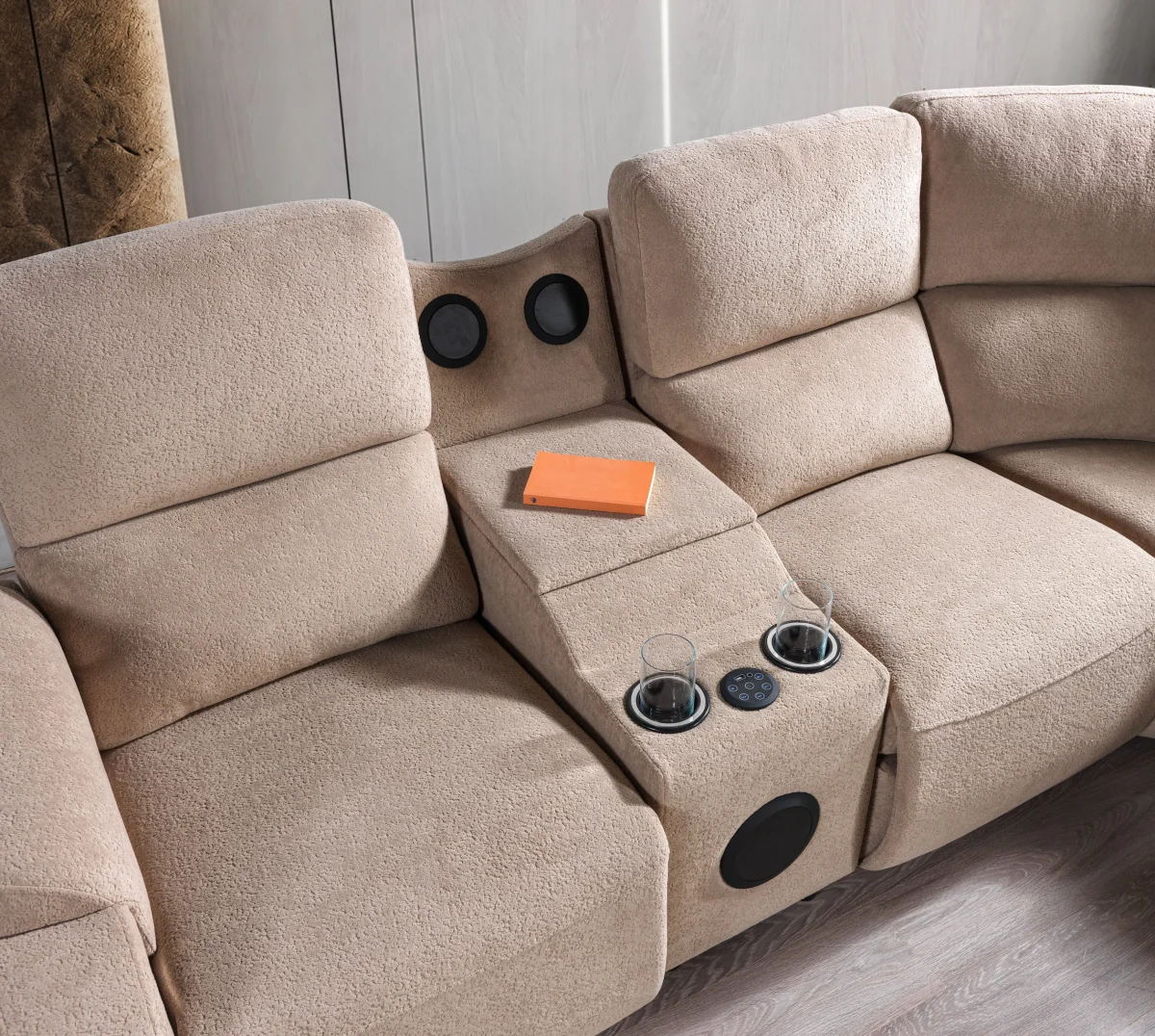 A detailed view of the Atlas Recliner Köşe Takımı, a contemporary L-shaped corner sectional sofa upholstered in textured beige fabric. The image shows its modern design with sleek lines and wide seats. The central console unit is featured, with an open lid, integrated cup holders, and a built-in Bluetooth speaker system visible on the side. The sofa is placed in a premium living room with light wood floors and textured wall paneling with LED backlighting.