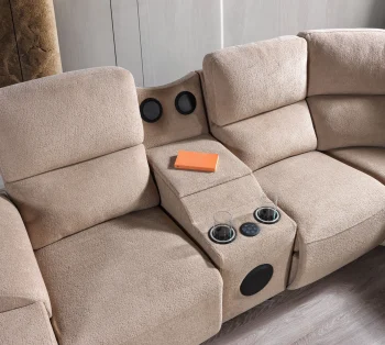 A detailed view of the Atlas Recliner Köşe Takımı, a contemporary L-shaped corner sectional sofa upholstered in textured beige fabric. The image shows its modern design with sleek lines and wide seats. The central console unit is featured, with an open lid, integrated cup holders, and a built-in Bluetooth speaker system visible on the side. The sofa is placed in a premium living room with light wood floors and textured wall paneling with LED backlighting.