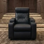 A front-facing view of the Zero Plus recliner, featuring a plush black leather design with geometric backrest stitching and built-in armrest cup holders, positioned in a sophisticated home cinema setting with illuminated, carpeted steps.