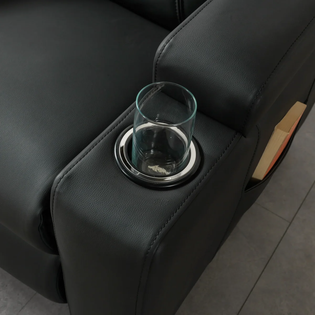 A front-facing view of the ZERO PLUS recliner, a luxurious black leather recliner chair with ergonomic stitched paneling and built-in cup holders. The recliner is centered on a light rug in a modern living room setting featuring elegant wood wall panels, accompanied by a tall woven vase holding decorative dry grass.